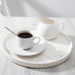 A small ceramic cup with coffe and milk jug for milk on a white tray.