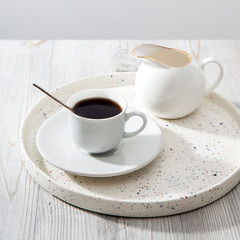 A small ceramic cup with coffe and milk jug for milk on a white tray.