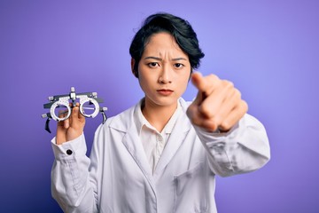 Beautiful asian optical girl wearing coat holding optometry glasses over purple background pointing with finger to the camera and to you, hand sign, positive and confident gesture from the front