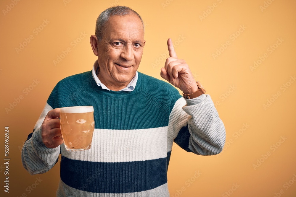 Sticker Senior handsome man drinking jar of beer standing over isolated yellow background smiling amazed and surprised and pointing up with fingers and raised arms.