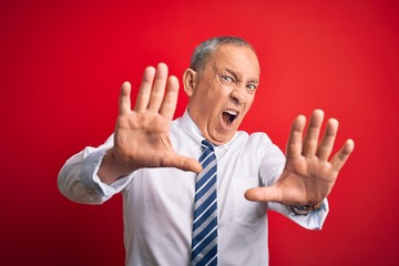 Senior handsome businessman wearing elegant tie standing over isolated red background afraid and terrified with fear expression stop gesture with hands, shouting in shock. Panic concept.