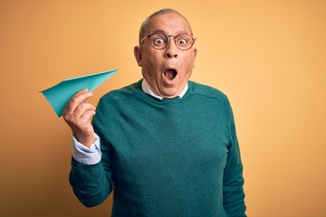 Senior handsome man holding airplane of paper standing over isolated yellow background scared in shock with a surprise face, afraid and excited with fear expression