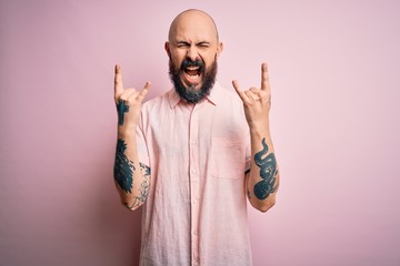 Handsome bald man with beard and tattoo wearing casual shirt over isolated pink background shouting...