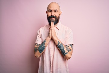 Handsome bald man with beard and tattoo wearing casual shirt over isolated pink background praying with hands together asking for forgiveness smiling confident.