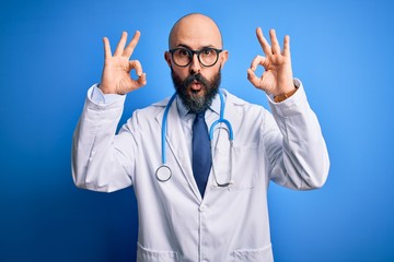 Handsome bald doctor man with beard wearing glasses and stethoscope over blue background looking surprised and shocked doing ok approval symbol with fingers. Crazy expression