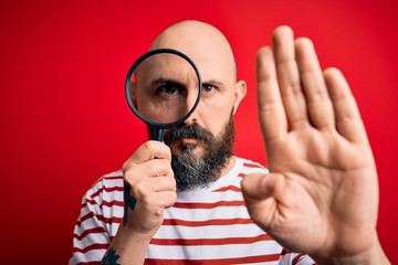 Handsome detective bald man with beard using magnifying glass over red background with open hand...