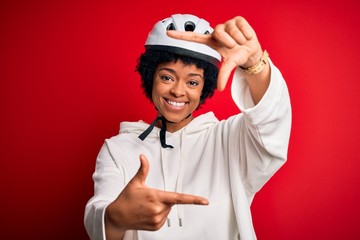 Young African American afro cyciling woman with curly hair wearing bike security helmet smiling making frame with hands and fingers with happy face. Creativity and photography concept.