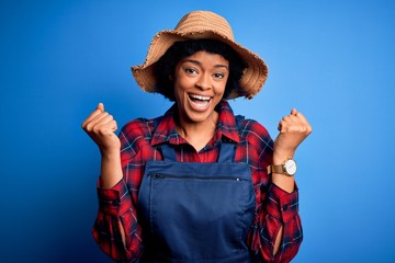 Young African American afro farmer woman with curly hair wearing apron and hat celebrating surprised and amazed for success with arms raised and open eyes. Winner concept.