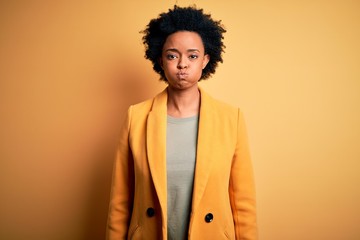 Fototapeta na wymiar Young beautiful African American afro businesswoman with curly hair wearing yellow jacket puffing cheeks with funny face. Mouth inflated with air, crazy expression.