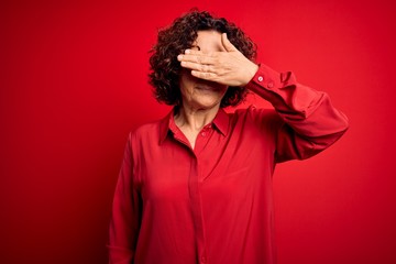 Obraz na płótnie Canvas Middle age beautiful curly hair woman wearing casual shirt and glasses over red background covering eyes with hand, looking serious and sad. Sightless, hiding and rejection concept