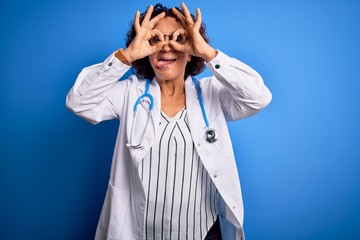 Middle age curly hair doctor woman wearing coat and stethoscope over blue background doing ok gesture like binoculars sticking tongue out, eyes looking through fingers. Crazy expression.