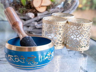 blue and golden indian singing bowl made of seven metals with a wooden striker on a glass table, plants in background