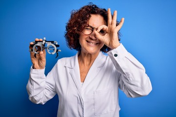 Middle age optical woman wearing coat holding optometry glasses over blue background with happy face smiling doing ok sign with hand on eye looking through fingers