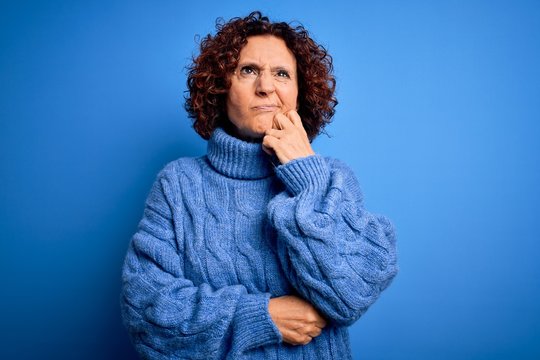 Middle age beautiful curly hair woman wearing casual turtleneck sweater over blue background with hand on chin thinking about question, pensive expression. Smiling with thoughtful face. Doubt concept.