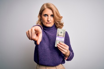 Middle age beautiful blonde woman holding bunch of dollars banknotes over white background pointing with finger to the camera and to you, hand sign, positive and confident gesture from the front