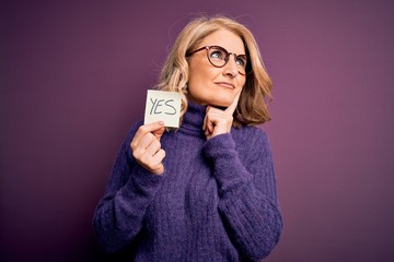 Middle age beautiful blonde woman wearing glasses holding reminder paper with yes message serious...
