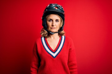 Middle age beautiful blonde motorcyclist woman wearing moto helmet over red background with serious expression on face. Simple and natural looking at the camera.