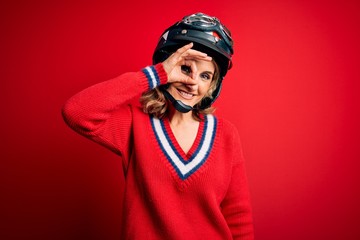 Middle age beautiful blonde motorcyclist woman wearing moto helmet over red background doing ok gesture with hand smiling, eye looking through fingers with happy face.