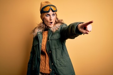 Middle age beautiful blonde skier woman wearing snow sportwear and ski goggles Pointing with finger surprised ahead, open mouth amazed expression, something on the front