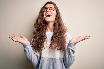 Young beautiful woman with curly hair wearing sweater and glasses over white background celebrating...