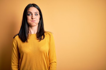 Young brunette woman with blue eyes wearing casual sweater over yellow background puffing cheeks with funny face. Mouth inflated with air, crazy expression.