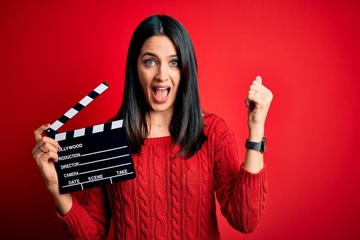 Young director woman with blue eyes making movie holding clapboard over red background screaming...