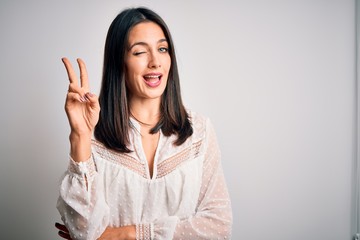 Young brunette woman with blue eyes wearing casual t-shirt over isolated white background smiling with happy face winking at the camera doing victory sign. Number two.