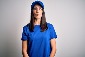 Young delivery woman with blue eyes wearing cap standing over blue background making fish face with lips, crazy and comical gesture. Funny expression.