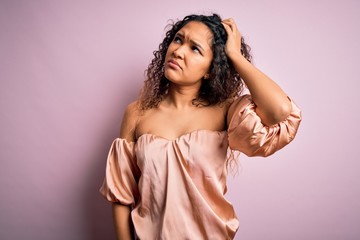 Young beautiful woman with curly hair wearing casual t-shirt standing over pink background confuse...