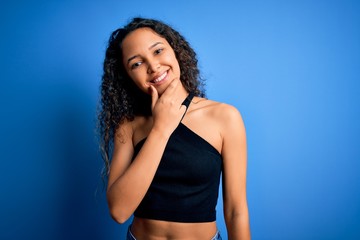 Young beautiful woman with curly hair wearing casual t-shirt standing over blue background looking confident at the camera smiling with crossed arms and hand raised on chin. Thinking positive.