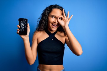 Young beautiful woman with curly hair holding broken smartphone showing cracked screen with happy face smiling doing ok sign with hand on eye looking through fingers