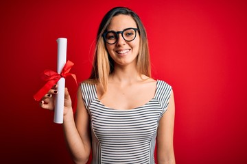 Young blonde woman with blue eyes wearing glasses holding graduated degree diploma with a happy face standing and smiling with a confident smile showing teeth