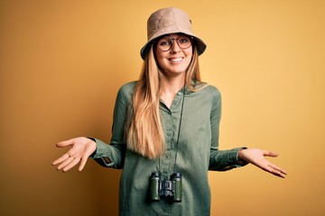 Beautiful blonde explorer woman with blue eyes wearing hat and glasses using binoculars clueless...