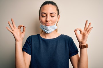 Beautiful blonde woman with blue eyes wearing medical mask over white background relax and smiling with eyes closed doing meditation gesture with fingers. Yoga concept.