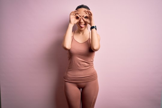 Young blonde fitness woman wearing sport workout clothes over pink isolated background doing ok gesture like binoculars sticking tongue out, eyes looking through fingers. Crazy expression.