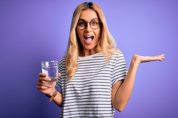 Young blonde healthy woman wearing glasses drinking glass of water over purple background very happy and excited, winner expression celebrating victory screaming with big smile and raised hands