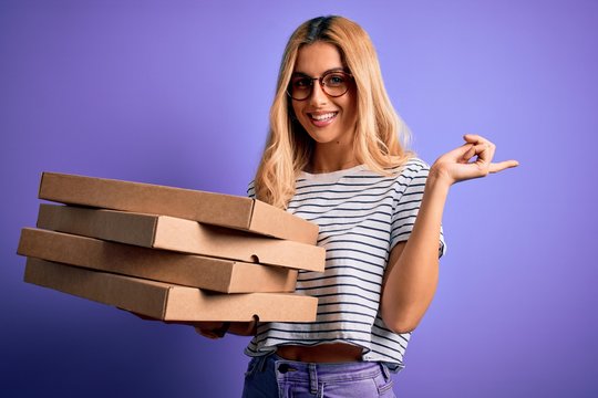 Young beautiful blonde woman holding boxes of italian pizza over isolated purple background very happy pointing with hand and finger to the side