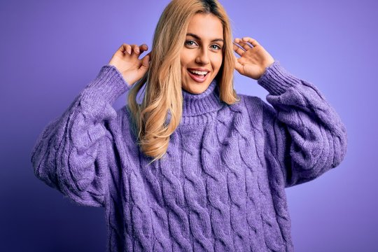 Young beautiful blonde woman wearing casual turtleneck sweater over purple background Smiling pulling ears with fingers, funny gesture. Audition problem