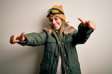 Young brunette skier woman wearing snow clothes and ski goggles over white background approving doing positive gesture with hand, thumbs up smiling and happy for success. Winner gesture.