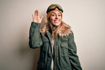 Young brunette skier woman wearing snow clothes and ski goggles over white background Waiving saying hello happy and smiling, friendly welcome gesture