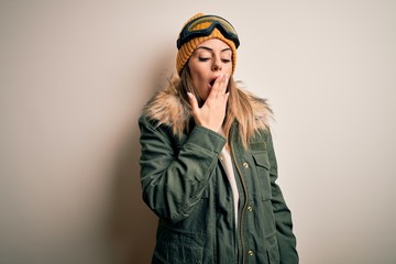 Young brunette skier woman wearing snow clothes and ski goggles over white background bored yawning tired covering mouth with hand. Restless and sleepiness.