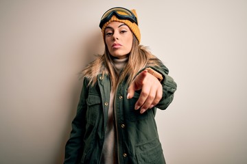 Young brunette skier woman wearing snow clothes and ski goggles over white background pointing displeased and frustrated to the camera, angry and furious with you