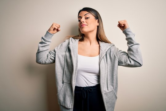 Young beautiful brunette sportswoman wearing sportswoman training over white background showing arms muscles smiling proud. Fitness concept.