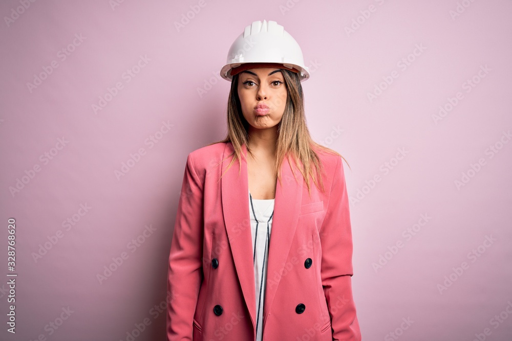 Sticker young beautiful brunette architect woman wearing safety helmet over pink background puffing cheeks w