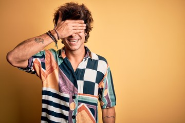 Young handsome man on vacation wearing summer shirt over isolated yellow background smiling and laughing with hand on face covering eyes for surprise. Blind concept.