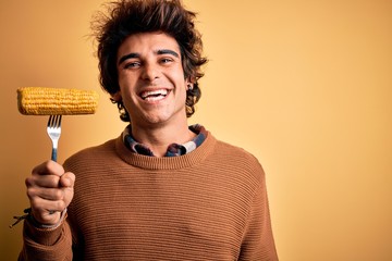 Young handsome man holding fork with cob corn standing over isolated yellow background with a happy face standing and smiling with a confident smile showing teeth
