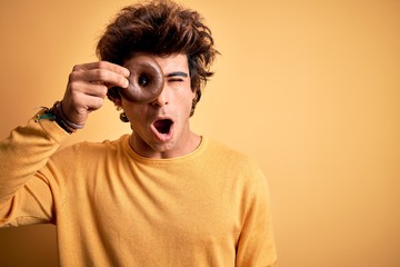 Young handsome man holding chocolate donut standing over isolated yellow background scared in shock with a surprise face, afraid and excited with fear expression