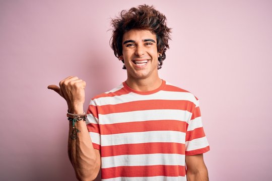 Young handsome man wearing striped casual t-shirt standing over isolated pink background smiling with happy face looking and pointing to the side with thumb up.