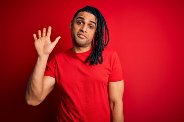 Young handsome african american afro man with dreadlocks wearing red casual t-shirt Waiving saying hello happy and smiling, friendly welcome gesture