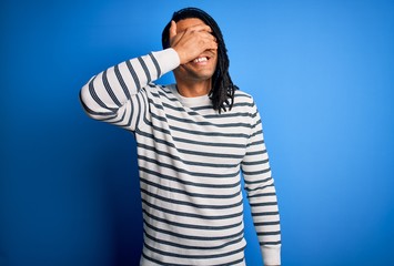 Young handsome african american afro man with dreadlocks wearing casual striped sweater smiling and laughing with hand on face covering eyes for surprise. Blind concept.